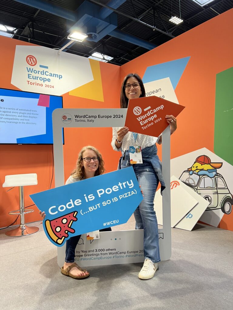 Two women are posing inside a large photo frame prop at WordCamp Europe 2024 in Torino, Italy. The woman on the left is crouching and holding a blue sign that reads "Code is Poetry (...BUT SO IS PIZZA) #WCEU" with a slice of pizza graphic. The woman on the right is standing and holding an orange arrow-shaped sign that says "WordCamp Europe Torino 2024." The background features bright orange walls with colorful geometric shapes and WordCamp Europe branding. A cartoon dog wearing a red cap is illustrated on a white hexagonal panel to the right.