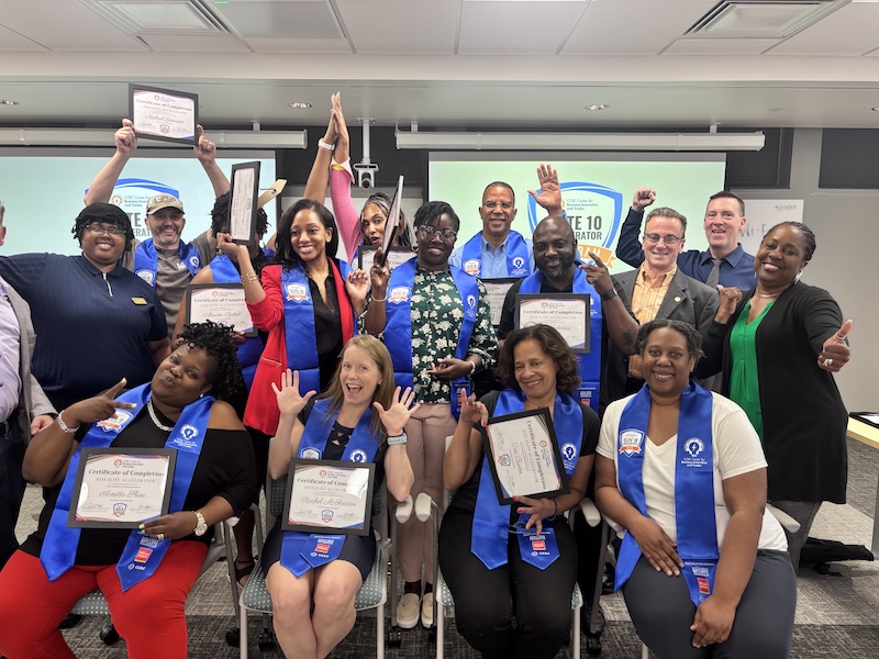 A group of diverse adults posing indoors, many wearing blue stoles and holding certificates of completion. They are smiling, raising their hands, and making celebratory gestures, indicating a graduation or achievement event. The background includes a projection screen with partially visible text and logos.