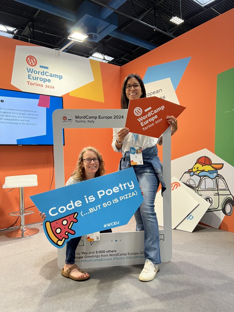 Two women posing at WordCamp Europe 2024 in Torino, Italy, holding a large photo frame prop and a blue sign that reads "Code is Poetry (...BUT SO IS PIZZA)" with a pizza slice illustration. The background features bright orange walls with WordCamp Europe branding and a cartoon image of a yellow dog wearing a red cap sitting on a car roof.
